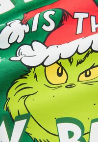 Groene stof met een cartoonachtige Grinch-gezicht, gele ogen en een witte hoedomtrek, met vetgedrukte tekst in wit en rood.