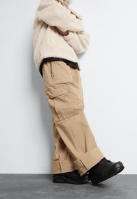 Person klädd i beige cargobyxor, svarta sneakers och en krämfärgad fleecejacka stående med armarna i kors mot en vit vägg.