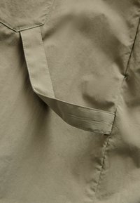 Hellbrauner Stoff mit einer gesteppten Klappe und Riemendetail, der die Textur und Nähte eines Kleidungsstücks zeigt.