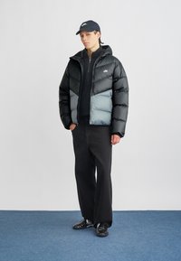 Mustan ja heloharmaan värinen puhku-takki, jossa on vetskari edessä, huppu ja Nike logo rinnassa, yhdistetty mustiin housuihin ja tennareihin.