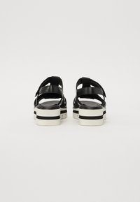 Sandales plateforme noires avec plusieurs brides croisées, présentant un dessus texturé et une semelle blanche avec des rayures noires. La vue de dos montre une boucle ajustable.
