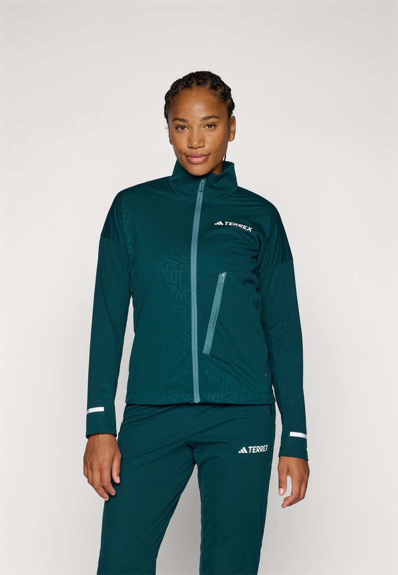 Veste de sport teal avec col haut, fermeture éclair et motif géométrique ton sur ton. Comprend une poche latérale et des accents réfléchissants.
