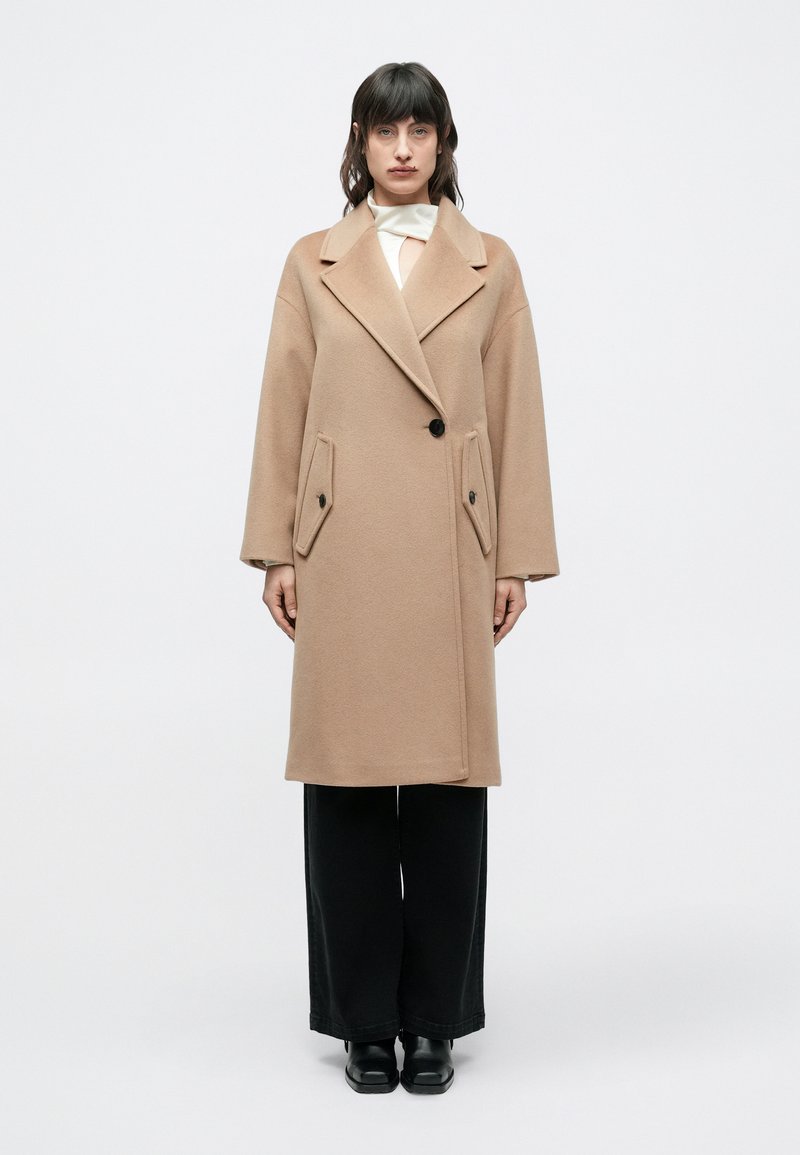 Bézs gyapjúkabát dupla soros gombolással, széles hajtókákkal, két első zsebbel és fekete gombokkal. Fekete, bő szárú nadrág fölött viselve.