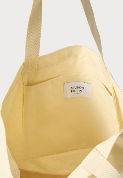 Interior de una bolsa de lona amarilla, con un bolsillo lateral y una etiqueta que dice "MAISON KITSUNÉ PARIS". Textura suave y correas resistentes.