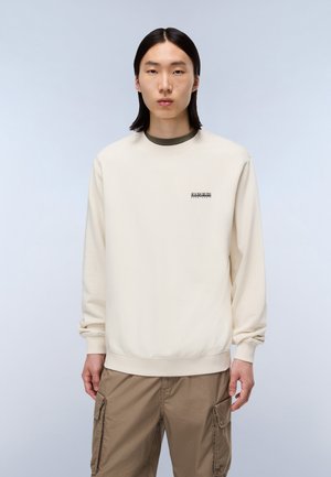 Jonge man met recht zwart haar, gekleed in een beige sweatshirt en bruine cargobroek, staande tegen een lichtgrijze achtergrond.
