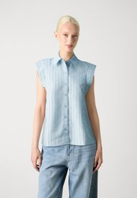Camicia senza maniche azzurro chiaro con righe verticali, chiusura con bottoni, colletto a punta e tessuto morbido. Indossata con jeans in denim larghi.