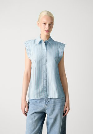 Camisa sin mangas de color azul claro con rayas verticales, con botones en la parte delantera, cuello puntiagudo y tejido suave. Llevada con unos vaqueros de corte holgado.