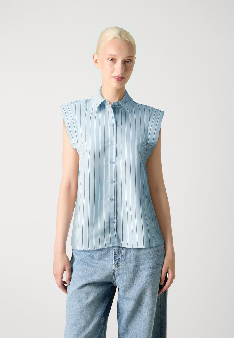 Camicia senza maniche azzurro chiaro con righe verticali, chiusura con bottoni, colletto a punta e tessuto morbido. Indossata con jeans in denim larghi.