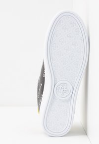 Baskets avec une semelle en caoutchouc blanc présentant un motif texturé. Le côté affiche un corps en tissu noir avec des logos "GUESS" blancs répétés.