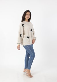 Maglione beige con motivi a stelle nere, texture a maglia e maniche a palloncino, abbinato a jeans skinny blu e sandali con tacco trasparente.
