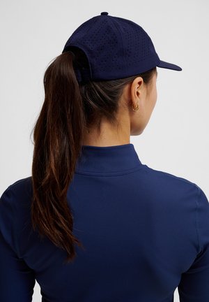 Casquette de baseball bleu marine avec un design perforé, sangle ajustable à l'arrière, portée avec un haut à manches longues bleu marine assorti.