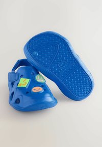 Zoccoli di plastica blu con fori di ventilazione, cinturino regolabile e charm decorativi colorati che presentano faccine sorridenti e forme sulla superficie superiore.