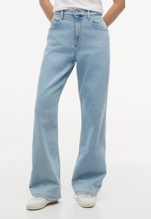 Ανοιχτό μπλε τζιν ψηλόμεσο με φαρδιά πόδια, φορεμένο με λευκά αθλητικά παπούτσια, εμφανίζεται σε ένα άτομο που στέκεται με τα χέρια πίσω από την πλάτη.