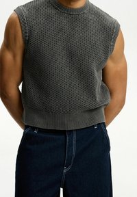 Grå stickad ärmlös tröja med en strukturerad mönster och ribbad rundhals, i kombination med mörka jeans med vit sömnad.