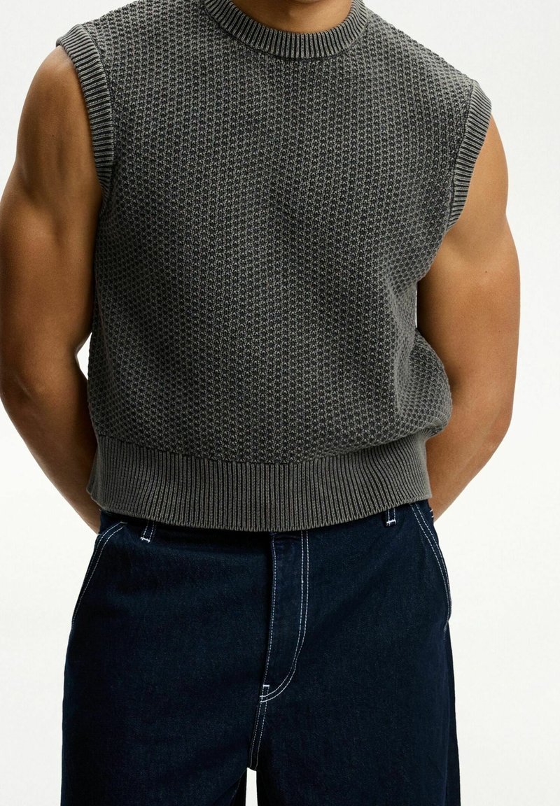 Grå stickad ärmlös tröja med en strukturerad mönster och ribbad rundhals, i kombination med mörka jeans med vit sömnad.
