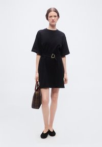 Γυναίκα στέκεται φορώντας κοντό μαύρο φόρεμα με ζώνη και μαύρα φλατ παπούτσια, κρατώντας καφέ καρό τσάντα, μπροστά σε απλό λευκό φόντο.