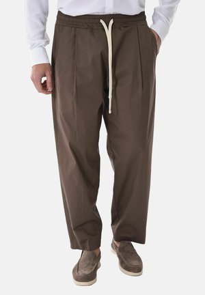 Uomo con pantaloni marroni larghi con coulisse bianca, camicia bianca e scarpe slip-on in camoscio marrone, in piedi davanti a uno sfondo semplice.