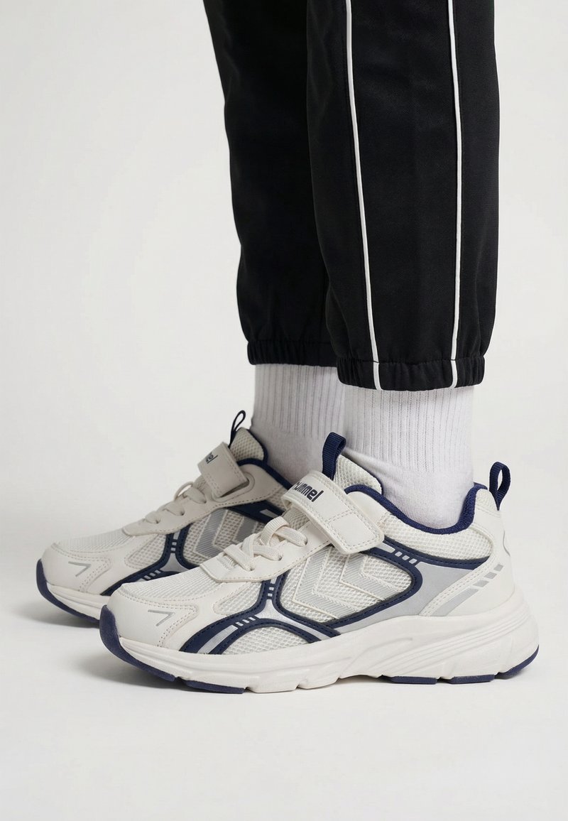 Kind draagt witte en marineblauwe ademende sneakers met klittenbandsluitingen, witte sokken en zwarte broek met witte zijstrepen.