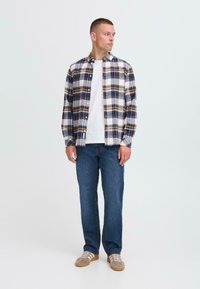 Karirana flanelasta srajca v modri, beli in rumeni barvi, gumbasta, v kombinaciji s svetlo modrimi jeans hlačami in sivimi supergami z belimi črtami.