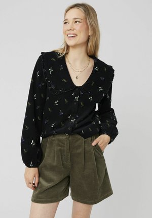 Chemise noire à fleurs avec col volanté, manches longues et fermeture à boutons, associée à un short en velours côtelé vert olive avec des plis.