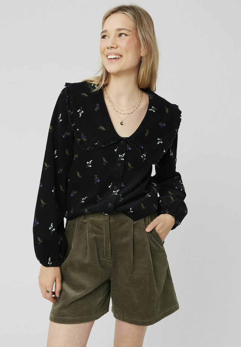 Chemise noire à fleurs avec col volanté, manches longues et fermeture à boutons, associée à un short en velours côtelé vert olive avec des plis.