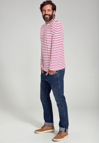 Rød og hvidstribet langærmet skjorte, denimjeans og lysebrune sneakers. Skjorten har en afslappet pasform; jeansene er tætsiddende med opfoldede manchetter.