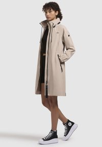 Trench coat impermeabile beige con colletto alto, dettagli in zip neri, polsini elasticizzati e cordoncino regolabile. Indossato con sneakers nere alte.