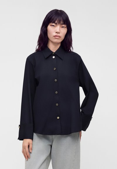 Femme portant une chemise noire à manches longues avec des boutons dorés et un jean gris clair sur un fond clair uni.