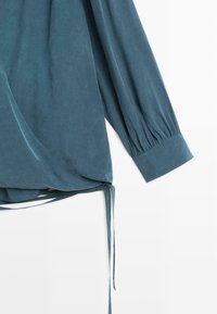 Blouse cache-cœur bleu sarcelle avec manches longues bouffantes et ourlet arrondi. Dotée d'un détail noué en bas et d'une texture lisse et douce.