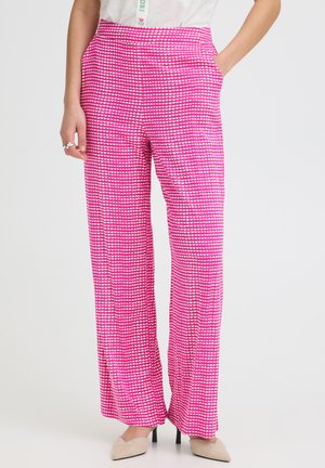 Femme portant un pantalon large rose vif avec de petits motifs de cœurs blancs, associée à des talons pointus beiges, debout les mains dans les poches.