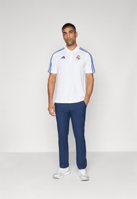 Polo bianco con accenti blu e un logo, abbinato a pantaloni blu scuro e sneakers chiare. Texture liscia, design del colletto standard.