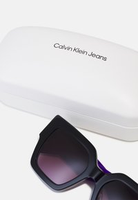 Mustad mustad prillid gradient tumedate läätsedega ja mati tekstuuriga, valge kõva kasti kõrval, millel on logo "Calvin Klein Jeans".