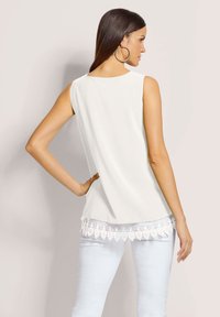 Weißes ärmelloses Oberteil mit Spitze am Saum, aus weichem Stoff und mit lockerer Passform. Kombiniert mit hellblauer Jeans.
