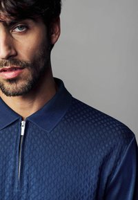 Polo de culoare albastru marin cu un model texturat în romburi, având un guler cu fermoar și o textură a materialului neted. Prim-plan al jumătății superioare a unui model.