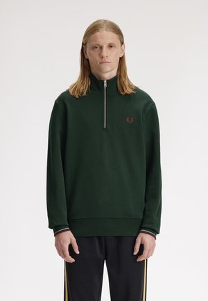 Jeune personne aux cheveux blonds mi-longs portant un sweat-shirt vert foncé à fermeture éclair quart et un pantalon noir avec des bandes jaunes sur les côtés.