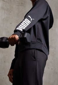 Sudadera atlética negra con el logo blanco de "PUMA" en la manga, combinada con pantalones negros; sosteniendo una mancuerna, tela texturizada, detalle de cremallera.