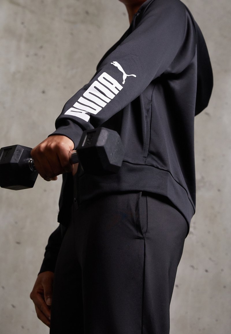 Sudadera atlética negra con el logo blanco de "PUMA" en la manga, combinada con pantalones negros; sosteniendo una mancuerna, tela texturizada, detalle de cremallera.