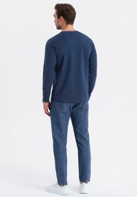 Uomo che indossa una camicia a maniche lunghe blu navy, jeans blu e sneakers bianche, in piedi di spalle contro uno sfondo chiaro e uniforme.