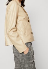Vrouw in een beige dubbelgestreepte neplederen jas met een hand in de zak van een grijs geruit broek, tegen een effen lichte achtergrond.