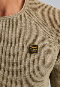 Beige gebreid sweater met geribbelde textuur, ronde hals en raglan mouwen. Voorzien van een zwart en gouden PME Legend merkpatch op de voorkant.
