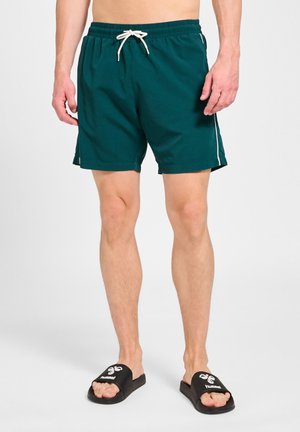Mand iført mørkegrønne badeshorts med hvid snøre og sorte slidesandaler med hvidt logo på en lys baggrund.