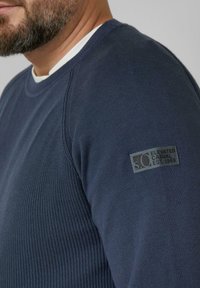 Marineblauer Strickpullover mit geripptem Muster, rundem Ausschnitt und einem Patch an der linken Ärmel mit Markendetails.