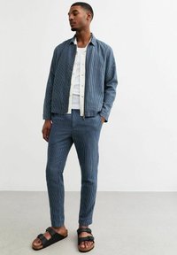 Blauwe gestreepte jas met bijpassende broek, van katoen, met een losse pasvorm, en zwarte sandalen met een kurken voetbed. Eenvoudig wit T-shirt eronder.