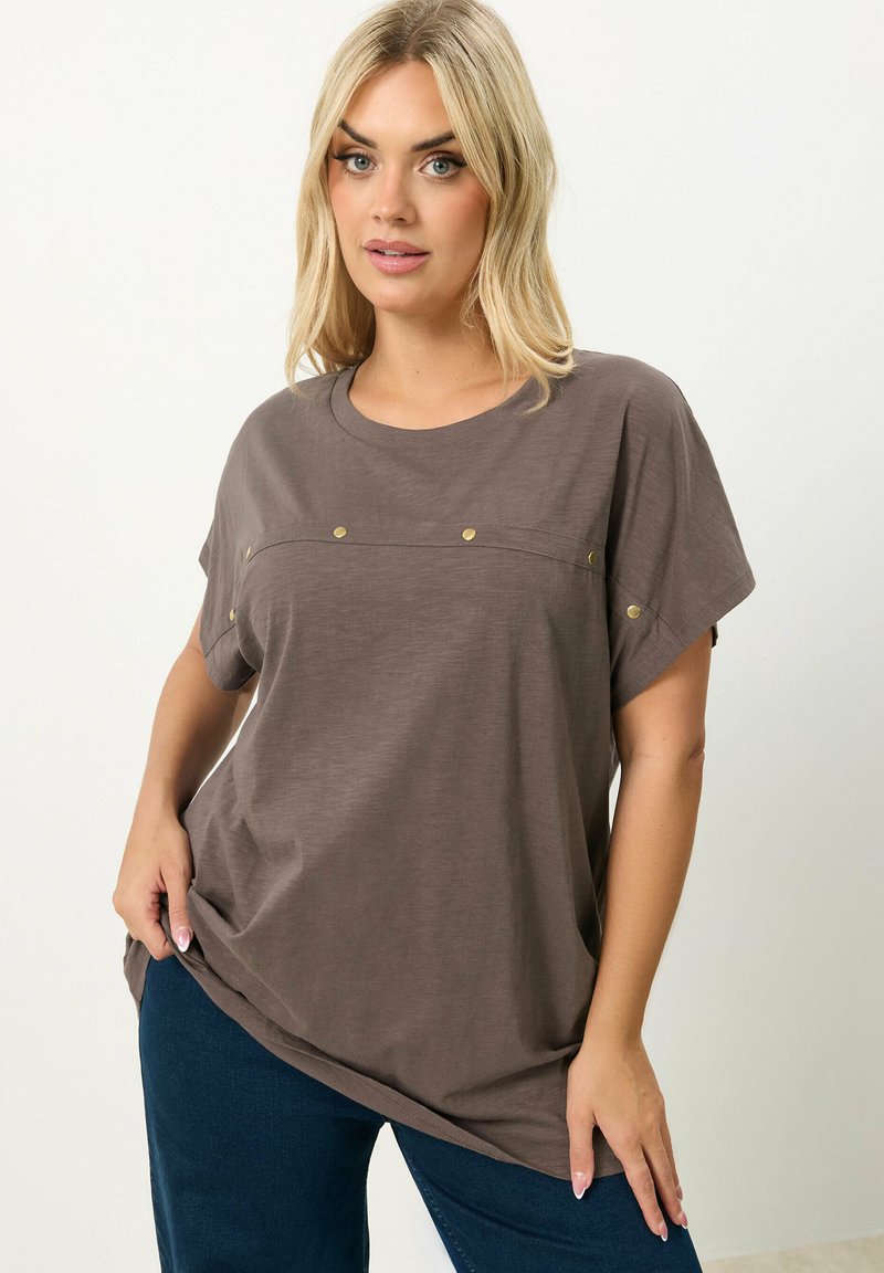 Blonde vrouw die een loszittend bruin shirt met korte mouwen draagt, met gouden knopen over de borst en mouwen, gecombineerd met donkerblauwe broek.