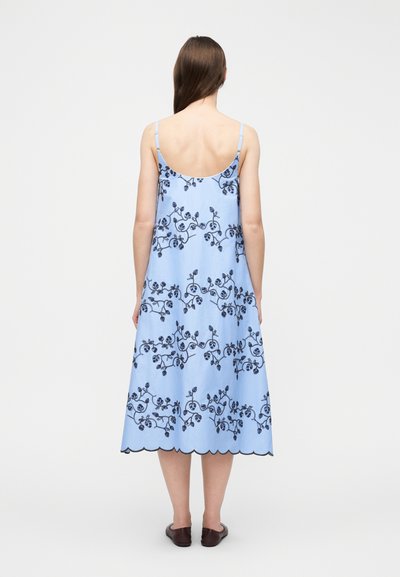 Γυναίκα με μακριά καστανά μαλλιά που φοράει ένα ανοιχτό μπλε μίντι φόρεμα με σκούρες ανθισμένες κεντήματα και τελείωμα με κυματιστό μοτίβο, στέκεται με την πλάτη σε λευκό φόντο.