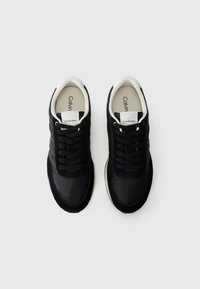 Zwarte sportieve schoenen met een gladde stoffen bovenkant, suède accenten, een contrasterende witte zool en zwarte veters. Merkenlabel op de tong.