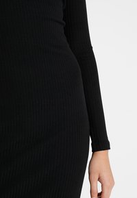 Černé žebrované šaty vyrobené z hladké látky, se dlouhými rukávy a přiléhavým střihem. Textura je jednotná po celé ploše oděvu.