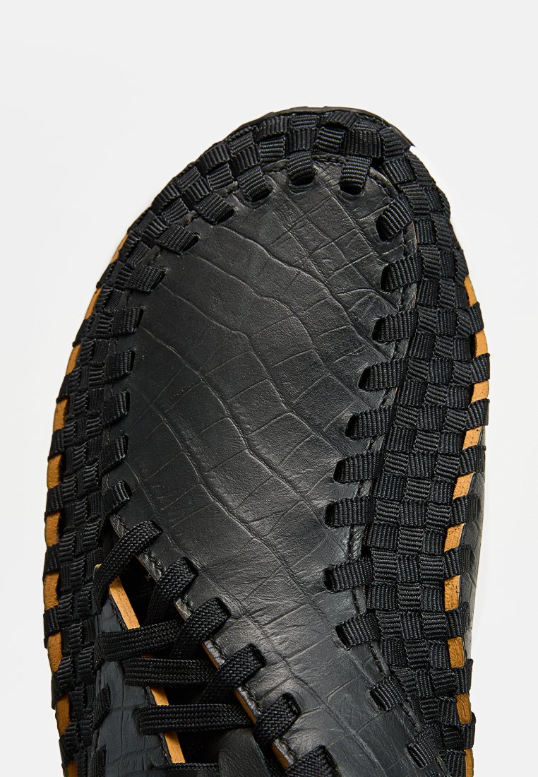 Chaussure en cuir noir avec une surface texturée, dotée de lanières tissées noires, d'accents jaunes et d'un design à bout rond.
