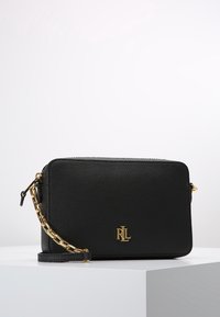 Černá kožená crossbody taška s texturovaným povrchem, zlatými kovovými detaily a řetízkovým popruhem. Na přední straně se nachází vyražené logo.