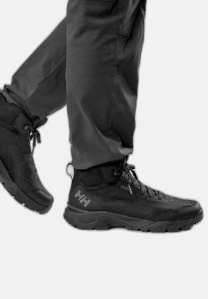 Persona che indossa stivali da trekking Helly Hansen neri con lacci, abbinati a pantaloni grigio scuro su uno sfondo chiaro e neutro.